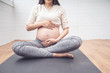 Body shot of pregnant woman caressing her baby belly, sitting on black yoga mat training yoga meditation, healthy and fit motherhood feeling happy and strong, pregnancy expecting future new born
