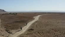 A Low Aerial POI Pullback Of Two Cyclists Bikepacking Along A Narrow Gravel Road Cutting Through A Barren Desert Landscape, With Small Buildings And A Dark Mountain Ridge In The Distance.