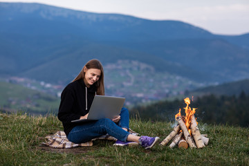 young female freelancer working on laptop in the mountains in the evening. Tourist girl sitting near campfire and having fun. Copy space.