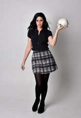 Wall Mural -  Portrait of a goth girl with dark hair wearing blue and plaid skirt with boots. Full length standing pose, holding a human skull, on a studio background.