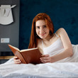 © Dmitrii Kotin - Redhead woman lying and relax on the bed and looks in her diary.