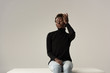 tender african american girl in jeans and black turtleneck sitting on white cube isolated on grey