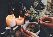 Female wiccan witch holding grey clay pot in her hands preparing ingredients for a spell at her altar. Dried plants, herbs, flowers, lit burning candles, black mirror and old bottles in the background