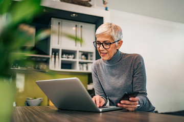 Wall Mural - Modern mature woman using laptop and smart phone at home, portrait.