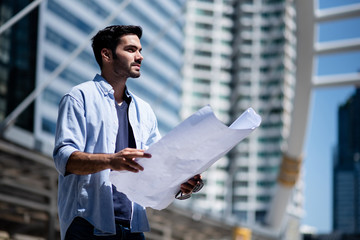 Young engineer in casual stand in city building background holding his blueprint and look forward to his work. Engineering construction, contractor business, new construction plan project concept.