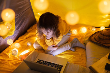 Wall Mural - young girl student using laptop for homeschooling in a home made tent with glowing light balls.