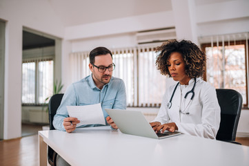 Female doctor, resident, using a laptop while sitting at desk with mentor who looking and holding paper document.