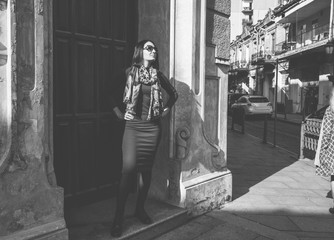 Wall Mural - Brunette woman with a positive smile in a dress and leather black jacket walks the street and enjoys a sunny spring day. Girl travel in Batumi, Georgia. Old city at background.