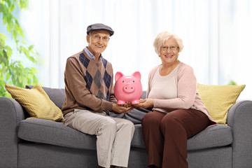 Wall Mural - Elderly couple on a sofa at home holding a piggy bank