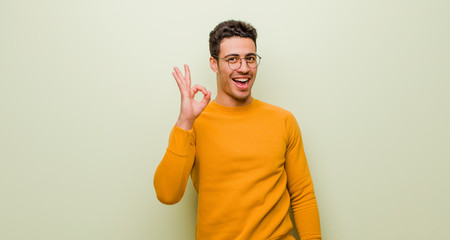 young arabian man feeling successful and satisfied, smiling with mouth wide open, making okay sign with hand against flat wall