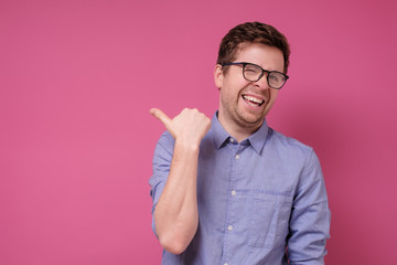 Wall Mural - Young man in glasses pointing with thumb finger aside isolated on pink background.