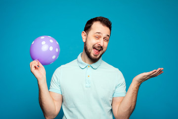 A handsome man holds a balloon in his hand and rejoices and screams at a holiday or birthday. A man inhaled laughing gas, helium and laughs on a blue background in the studio