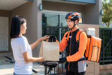 Asian man courier on bicycle delivering food in orange uniform smile and holding food bag in front house and Asian woman accepting a delivery of boxes from deliveryman.