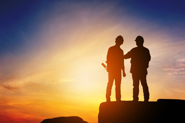 Silhouette of Engineer and worker standing on stone thinking of project sunset in evening time.