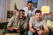 friendship, technology and leisure concept - smiling male friends with gamepads and beer playing video game at home at night