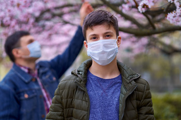 father and son with a face mask are in the city outdoor, blooming trees, spring season, flowering time - concept of allergies and health protection from dusty air