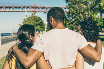 Wall Mural - Back view of friends embracing in park. Rear view of cheerful multiethnic friends hugging while walking together in park. Friendship concept