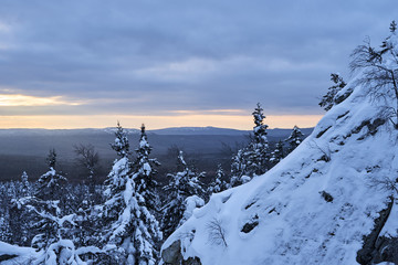 Winter forest with snow-covered fir trees high in the mountains. Dawn with bright colors on the horizon far away in the mountains. Golden clouds with the first rays of the sun.