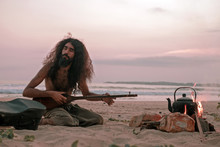A Man Plays A Stringed Ethnic Instrument On The Beach By The Fire And Heats Tea In A Teapot.