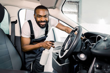 Close-up portrait of joyful smiling African male worker in uniform cleaning car interior and control panel with special cleaning chemical spray. Chemical and wet cleaning, car detailing concept