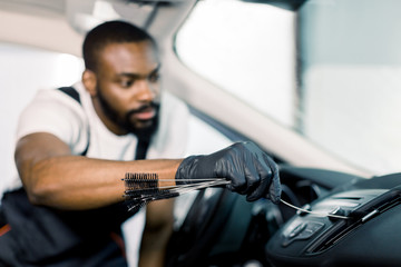 Young handsome concentrated African American man using cleaning brush and removing dust from car air conditioning vent grill. Car detailing or valeting concept. Focus on hand