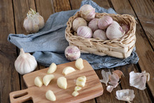 White Round Garlic Lies In A Wicker Basket On A Brown Wooden Background With A Blue Napkin. Next To The Cut Garlic Clove And A Knife. Close-up Photo