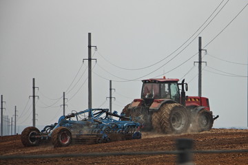 Red big twin wheels tractor with large plough ploughs the land in a field near the power lines masts poles at spring day, rural farm landscape