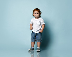 Little ginger toddler male or female in white t-shirt, socks and shoes, denim shorts. Child is smiling, posing on blue background