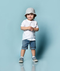 Ginger toddler boy or girl in white t-shirt, gray hat, socks and shoes, denim shorts. Child is smiling, posing on blue background