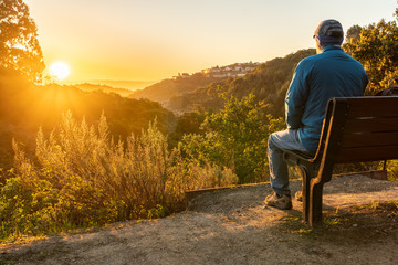 Person, man, sitting on a bench watching the sunrise over hills and valley with trees and shrubs in the middle and houses on a hill in the background
