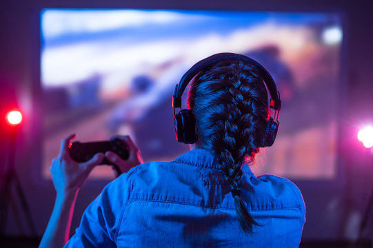 Young girl in headphones plays a video game on the big TV screen in the dark room. Gamer with a joystick. Online gaming at night, competitions, win, prize. Fun entertainment. Back view. Neon lighting
