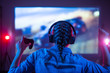 © Anton - Young girl in headphones plays a video game on the big TV screen in the dark room. Gamer with a joystick. Online gaming at night, competitions, win, prize. Back view. Neon lighting