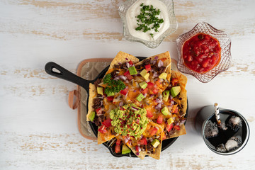 Wall Mural - Corn nachos chips topped with ground beef, melted cheddar cheese, and guacamole on white wooden background. Oven baked nachos in cast iron skillet with sour cream and salsa dips.