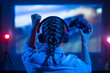 © Anton - Gamer plays a video game on a TV or PC screen. Girl with a joystick and headphones in a neon room. Online gaming, simulation games. Tournaments between players. Dark background, back view.