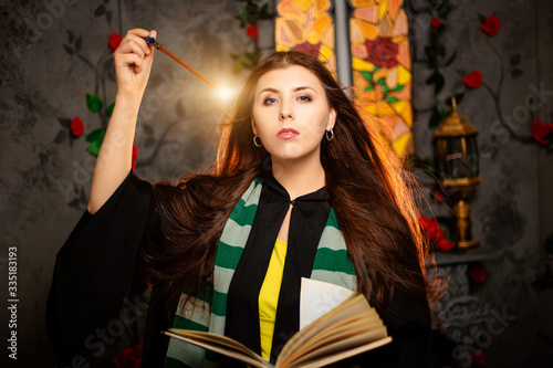 Fotomural A young woman in a black mantle with a striped scarf around her neck casts a spell from a book and conjures with a wand of magic
