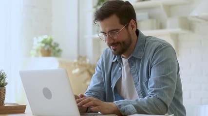 Concentrated young man freelancer in eyeglasses looking at laptop screen, working remotely at home. Head shot focused smiling guy chatting in social network, typing message, internet web surfing.