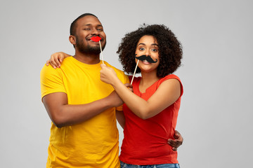 fun, photo booth and people concept - happy african american couple with party props hugging over grey background