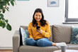 © Syda Productions - people, technology and leisure concept - happy smiling asian young woman in yellow sweater with smartphone sitting on sofa at home