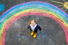 Happy Little Toddler Girl In Rubber Boots With Rainbow Painted With Colorful Chalks On Ground During Pandemic Coronavirus Quarantine. Children Painting Rainbows Along With The Words Let's All Be Well