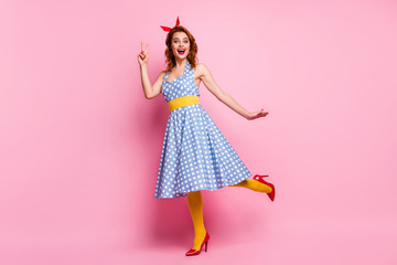 Full body photo of beautiful funny lady showing v-sign symbol stand on one leg wear headband dotted dress long skirt red stilettos yellow tights isolated pink color background