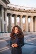 © KseniaJoyg - A young brunette girl with long hair in a jacket stands in the rays of the morning dawn on a deserted square in St. Petersburg against the background of the columns of the Kazan Cathedral