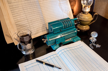 An old mechanical calculator, an arithmetic counter, accounting journals, a kerosene lamp, and a pen on a black table.