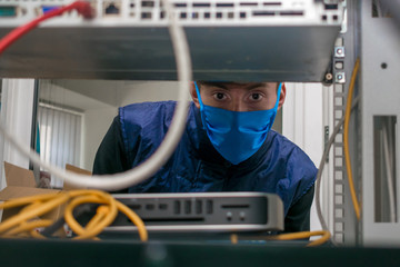 A technician in a medical mask works in a rack with computer equipment. The man serves the server room in the datacenter. Portrait of a teenager in a data center.