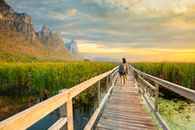 Asian Hikers Carry Heavy Backpacking On A Small Pavilion Outdoor Hiking Path On A Wooden Bridge In A Swamp With Meadows With A Blue Mountain Background. Khao Sam Roi Yot National Park. 
