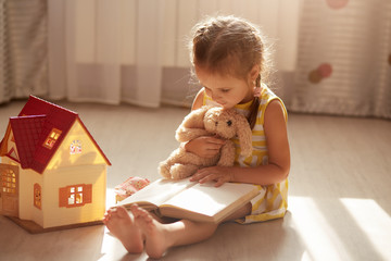 Little charming gir with pigtails reading her favorite book for her soft toy. Cute child reads stories, sitting on floor near toy house. Self isolation during coronavirus pandemic. Covid 19 concept.