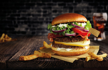 burger with double meat, cheese and french fries on a wooden table and parchment, with a flag, behind a black brick wall with takan cola