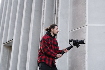 Wall Mural - Young Professional videographer holding professional camera on 3-axis gimbal stabilizer. Pro equipment helps to make high quality video without shaking. Cameraman wearing red shirt making a videos.