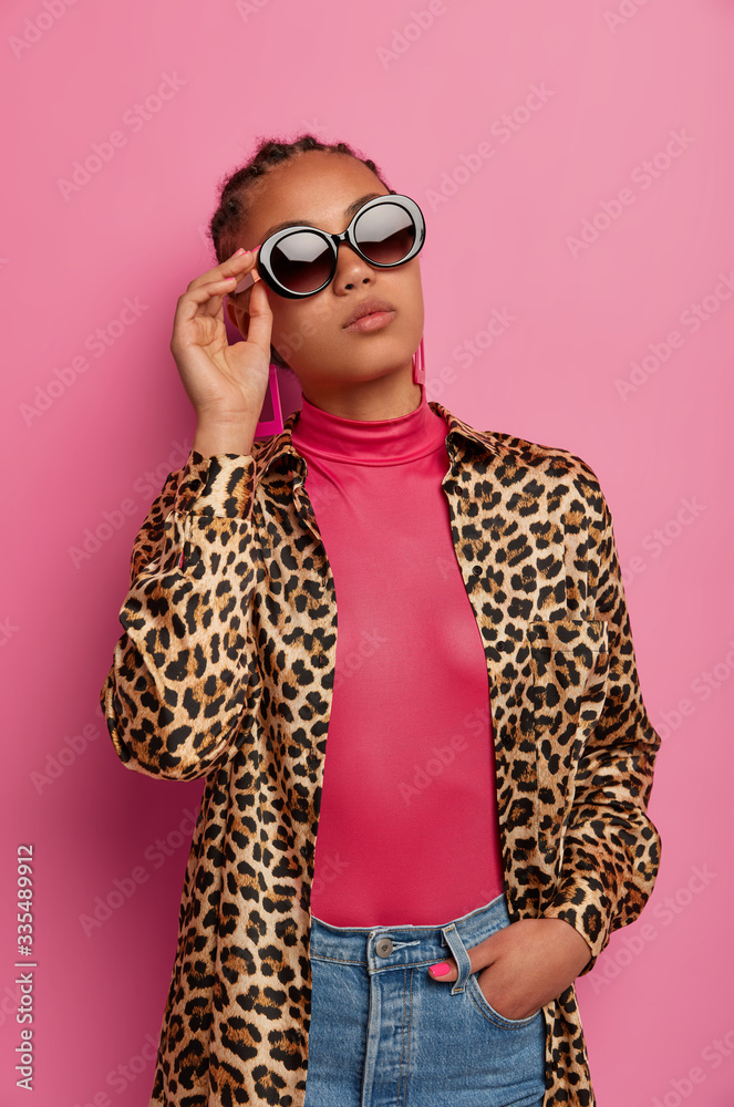 Photo of attractive fashionable woman wears sunglasses, shirt with leopard print, boasts of new purchases, being confident in herself, poses against pink background. Female in trendy clothes