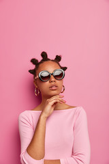 Beautiful dark skinned young woman keeps hand under chin, wears trendy summer sunglasses and stylish jumper, makes fashion photography, models in studio against pink background. Vogue, style