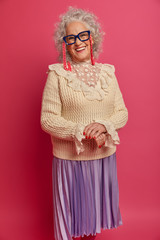 Positive wrinkled woman wears glasses, stylish clothes, smiles happily, expresses positive mood, being delighted or satisfied, isolated on pink background. Optimistic pensioner happy to retire